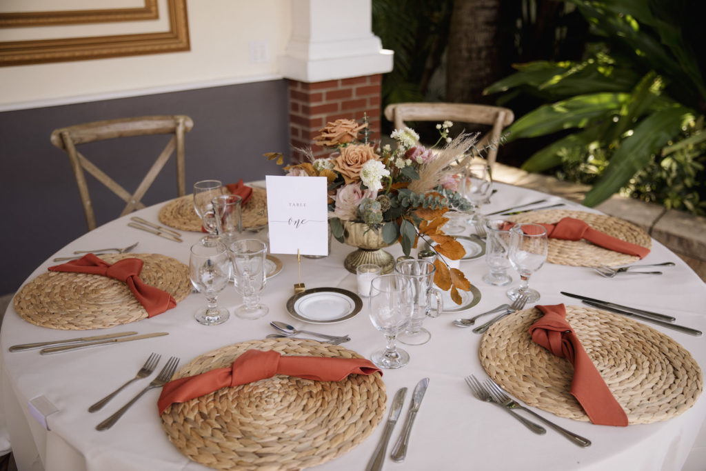 Wicker Place Mats with Burnt Orange Napkins on Cream Linen | Low Centerpieces with Roses, Greenery, and Pampas Leaves in Gold Holder | Bohemian Wedding Reception Decor | Sarasota Kate Ryan Event Rentals