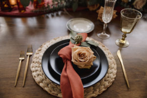 Wicker Placemat with Black Dishes Place Setting and Burnt Orange Napkins, Gold Flatware, and Rose Flower | Boho Wedding Reception Decor and Ideas