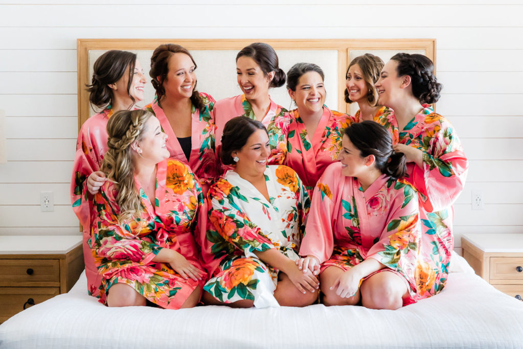Bride and bridesmaids in Floral Pink Matching Robes Sitting on Bed Getting Wedding Ready