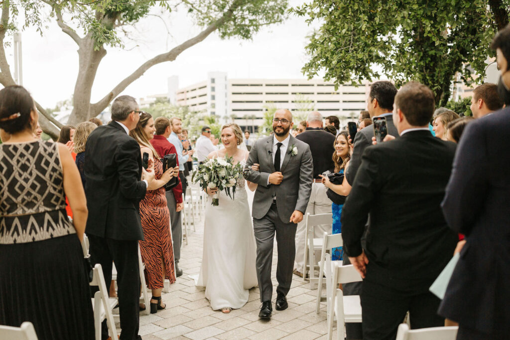 Bride and Groom Exit during Waterfront Outdoor Wedding Ceremony in Harbour Island Downtown Tampa | Groom Wearing Classic Charcoal Grey Suit