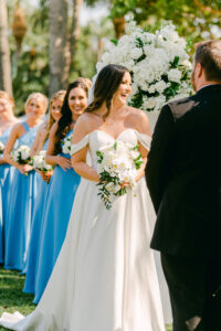 Florida Bride During Outdoor Ceremony, Wearing Off the Shoulder Stella York Dress, Holding White Cascading Floral Bouquet with Ivory Roses and White Orchids, Bridesmaids Wearing Slate Blue Long Dresses | Tampa Wedding Florist Brice Wayne Florals | Florida Wedding Planner Parties A La Carte