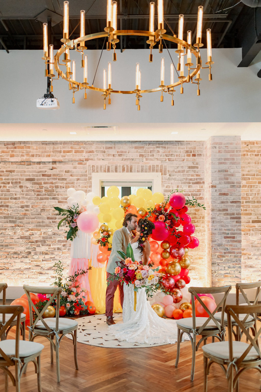 Bride and Groom Exchanging Wedding Vows, Whimsical and Colorful Wedding Decor, Yellow, Pink, Orange, White, Gold Fringe and Balloon Ceremony Backdrop, Wooden Cross Back Chairs, Gold and Candlestick Chandelier, White Brick Wall | Tampa Bay Wedding Photographer Dewitt for Love | St. Pete Modern Industrial Wedding Venue Red Mesa Events | Wedding Chair Rentals Kate Ryan Event Rentals