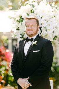Clearwater Groom During Wedding Ceremony, In front of towering floral agreement with white orchids, roses, and hydrangeas | Groom Wearing Classic Black Tuxedo with Bowtie | Florida Wedding Planner Parties A La Carte | Tampa Florist Bruce Wayne Florals