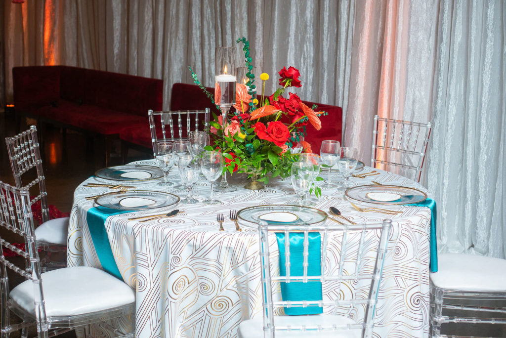 Modern Florida Wedding Reception Decor, Low Floral Centerpiece with Bright Red, Vibrant Orange and Tropical Greenery, White and Gold Art Deco Designed Linens, Gold Rimmed Crystal Charger with Teal Napkins and Gold FlatwareGhost Chiavari Chairs | 7th and Grove