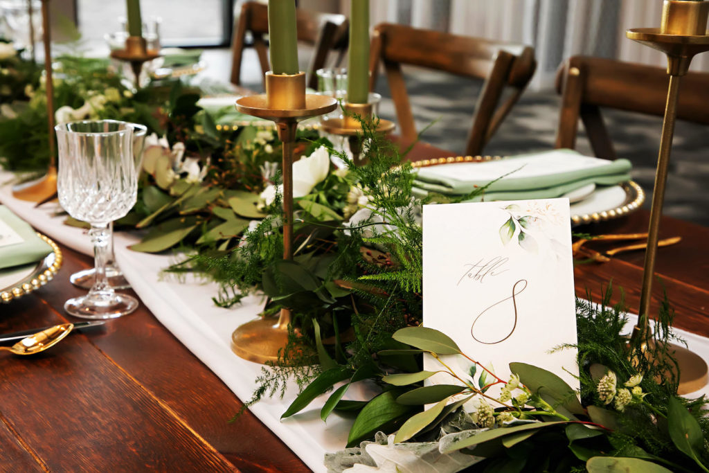 Romantic Wedding Reception Decor, Long Wooden Table with White Table Linen, Greenery Garland, Gold Candlesticks with Sage Green Candles, Gold and White Table Signage, Wooden Cross Back Chairs | Tampa Bay Wedding Photographer Limelight Photography | Wedding Planner MDP Events | Wedding Stationery A&P Design Co. | Wedding Florist Beneva Florals
