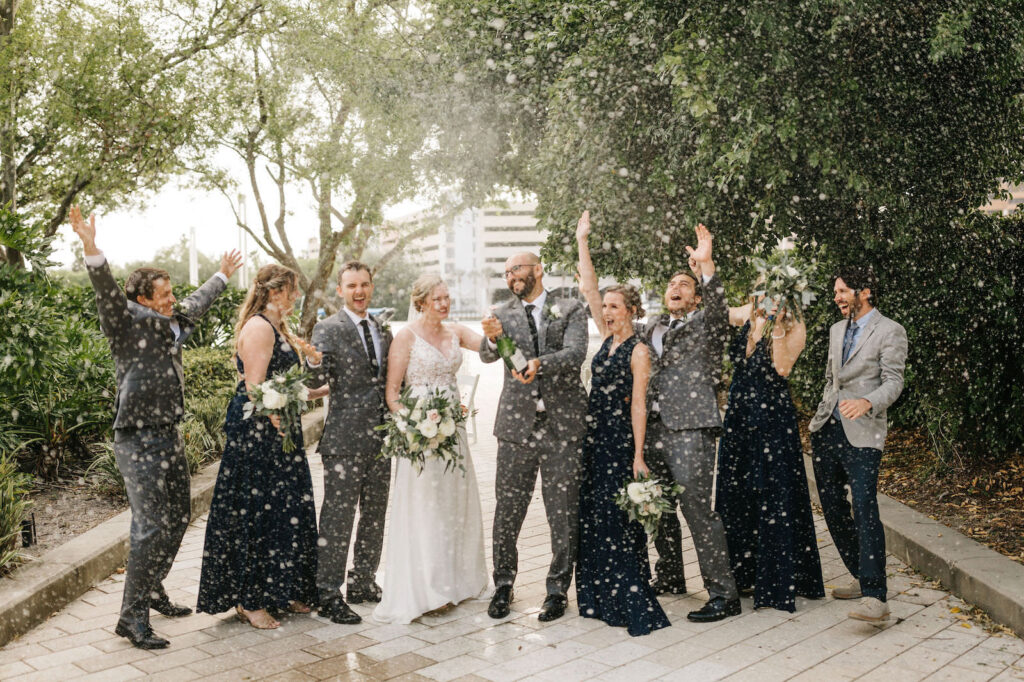 Bride and Groom Outdoor Wedding Party Portrait Champagne Pop Spray | Long Chiffon Navy Bridesmaid Dresses | Sheath Satin Charmeuse V Neck Lace Bodice Spaghetti Strap Bridal Gown | Groom and Groomsmen Wearing Classic Charcoal Grey Suit