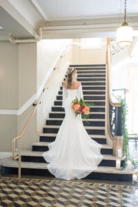 Florida Bride Wearing Vintage Lace and Fringe Off the Shoulder Sleeve Wedding Dress with Train and Gold, Flower Crown Holding Colorful Tropical Floral Bouquet Standing on Staircase, Pink Anthuriums, Monstera Palm Leaves | Tampa Bay Wedding Planner Eventfull Weddings | Historic Tampa Wedding Venue The Cuban Club