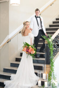 Florida Groom Wearing White and Black Collar Tuxedo, Bride Wearing Vintage Lace and Fringe Off the Shoulder Sleeve Wedding Dress with Train and Gold, Flower Crown Holding Colorful Tropical Floral Bouquet Standing on Staircase, Pink Anthuriums, Monstera Palm Leaves | Tampa Bay Wedding Planner Eventfull Weddings | Historic Tampa Wedding Venue The Cuban Club | Wedding Hair and Makeup Adore Bridal Hair and Makeup