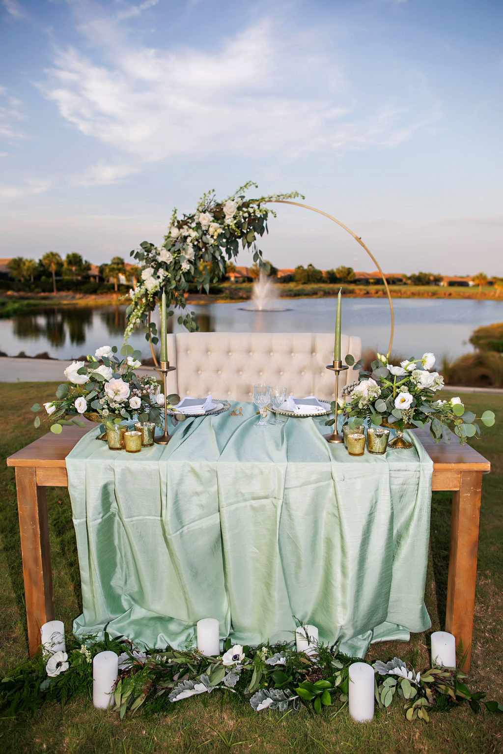 Outdoor Romantic Wedding Sweetheart Table Decor, Wooden Table, Ivory Tufted Loveseat, Gold Round Metal Arch with Floral Arrangement, White Anemone and Roses, Greenery, Eucalyptus, Sage Green Table Linen, Gold Mercury Votives, Gold Candlesticks and Sage Green Candles | Tampa Bay Wedding Photographer Limelight Photography | Wedding Planner MDP Events | Wedding Florist Beneva Florals | Wedding Venue Esplanade Country Club