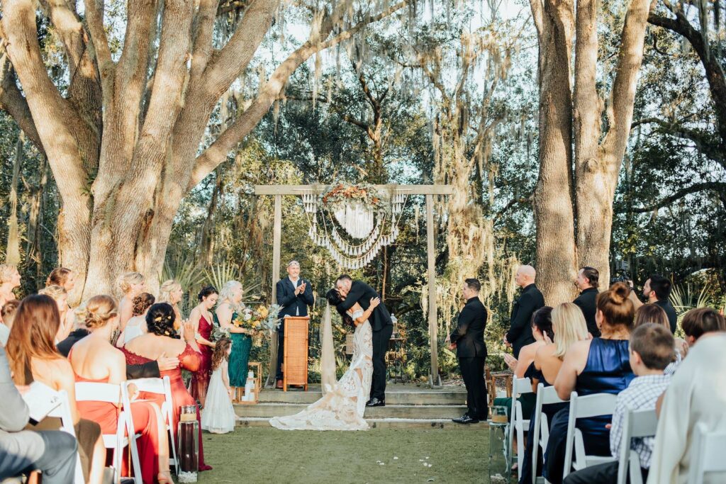 Bride and Groom First Kiss Portrait | Outdoor Bohemian Wedding Ceremony with White Garden Chairs | Wooden Arch with Crochet Details | Tampa Wedding Venue Paradise Springs
