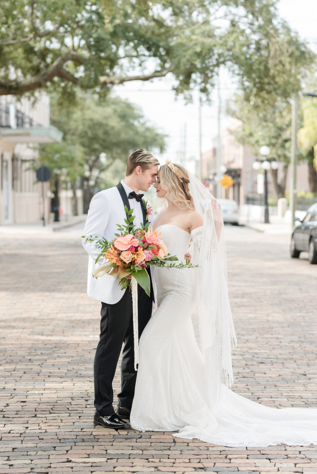 Vintage Bride Wearing Lace Strapless Sweetheart Neckline Fringe Off the Shoulder Sleeve Wedding Dress with Train, Gold Flower Crown Holding Colorful Tropical Floral Bouquet with Groom in White and Black Collar Tuxedo Standing in Streets of Tampa | Wedding Hair and Makeup Adore Bridal Hair and Makeup