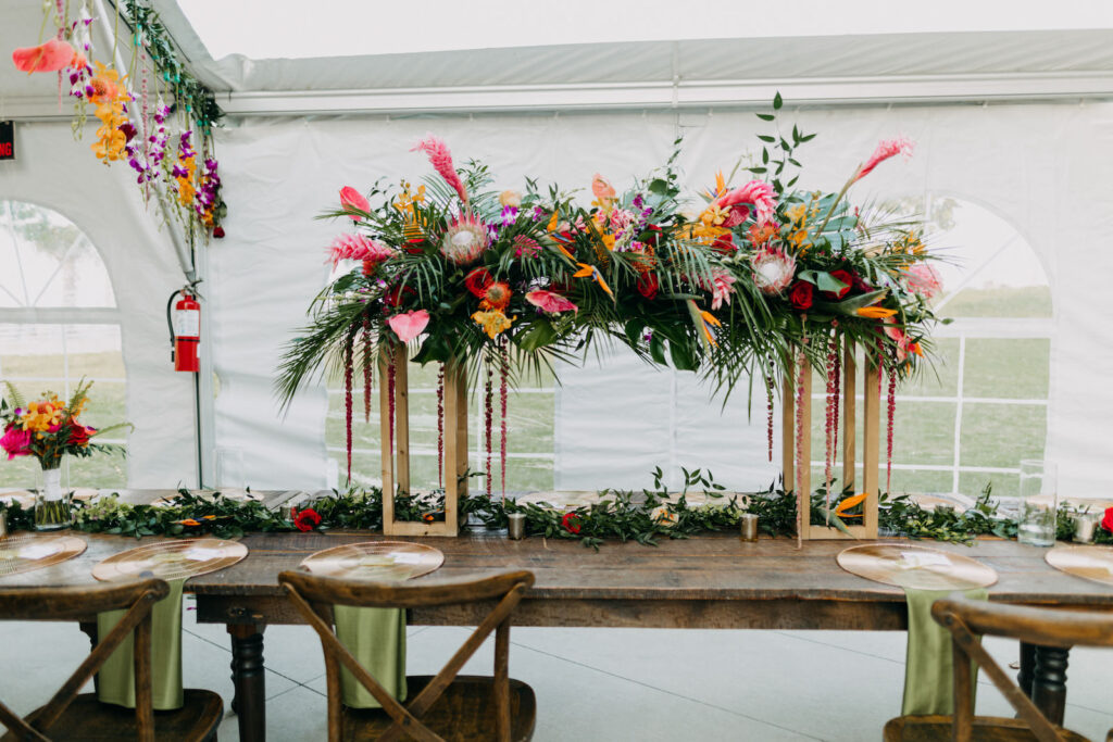 Tropical Boho Tent Wedding Reception Decor, Wooden Feasting Table with Wooden Crossback Chairs, Tall Rectangular Gold Stands with Palm Fronds, Monstera Leaves, Pink Ginger, Anthuriums, Orange Flowers, Pin Cushion Protea Lush Floral Centerpiece, Greenery Garland | Tampa Bay Wedding Photographer Amber McWhorter Photography | Wedding Rentals Kate Ryan Event Rentals