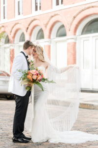 Vintage Bride Wearing Lace Strapless Sweetheart Neckline Fringe Off the Shoulder Sleeve Wedding Dress with Train, Gold Flower Crown and Fringe Veil Holding Colorful Tropical Floral Bouquet with Groom in White and Black Collar Tuxedo Standing in Streets of Tampa | Wedding Hair and Makeup Adore Bridal Hair and Makeup