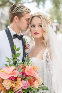 Vintage Bride Wearing Lace Strapless Sweetheart Neckline Fringe Off the Shoulder Sleeve Wedding Dress with Train, Gold Flower Crown and Fringe Veil Holding Colorful Tropical Floral Bouquet with Groom in White and Black Collar Tuxedo Standing in Streets of Tampa Portrait | Wedding Hair and Makeup Adore Bridal Hair and Makeup
