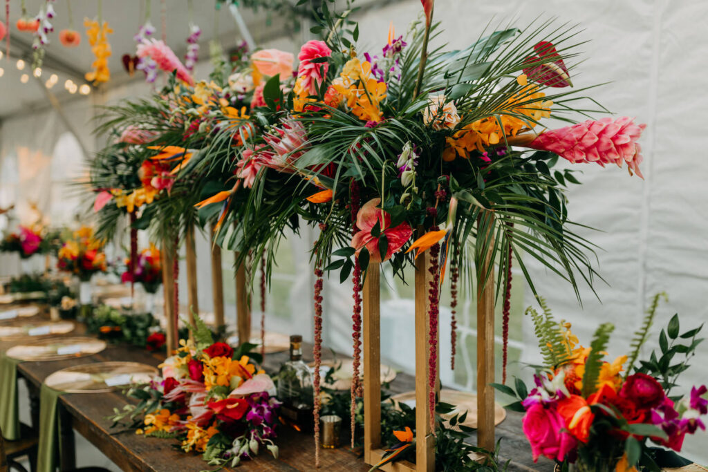 Tropical Boho Tent Wedding Reception Decor, Wooden Feasting Table with Wooden Crossback Chairs, Tall Rectangular Gold Stands with Palm Fronds, Monstera Leaves, Red Hanging Amaranthus, Pink Ginger, Anthuriums, Orange Flowers, Pin Cushion Protea Lush Floral Centerpiece, Greenery Garland | Tampa Bay Wedding Photographer Amber McWhorter Photography | Wedding Rentals Kate Ryan Event Rentals