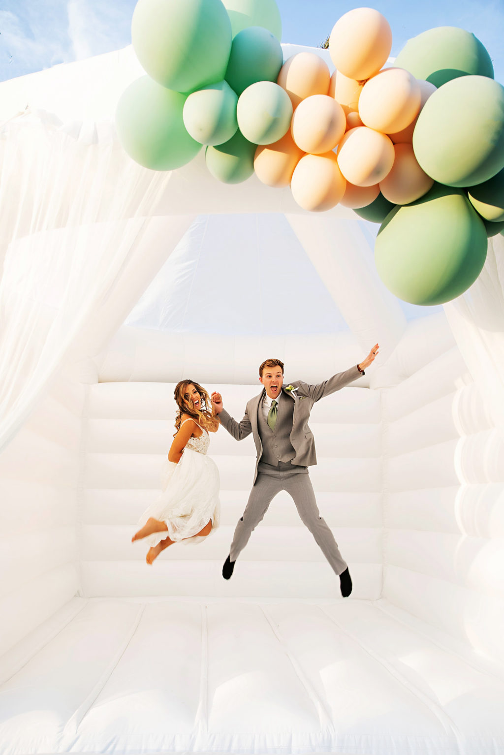 Bride and Groom Jumping on Fun, Unique White Adult Bounce House, Sage Green and Peach Balloons | Tampa Bay Wedding Photographer Limelight Photography | Wedding Planner MDP Events | Wedding Venue Esplanade Country Club | Wedding Dress Truly Forever Bridal Sarasota | Wedding Hair and Makeup Adore Bridal | Styled Shoot