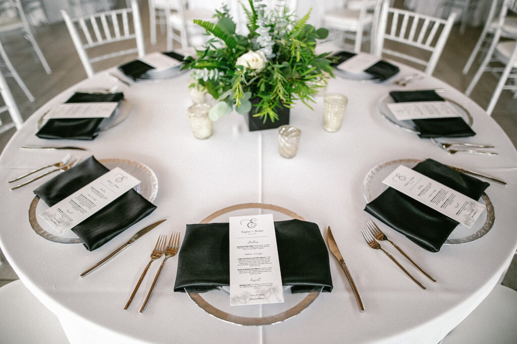 Tampa Modern Elegant Wedding Reception Decor, Clear and Silver Rimmed Chargers, Black Silk Napkins, Custom Menus, Low Floral Centerpiece with Greenery and White Flowers | Tampa Bay Wedding Rentals Kate Ryan Event Rentals