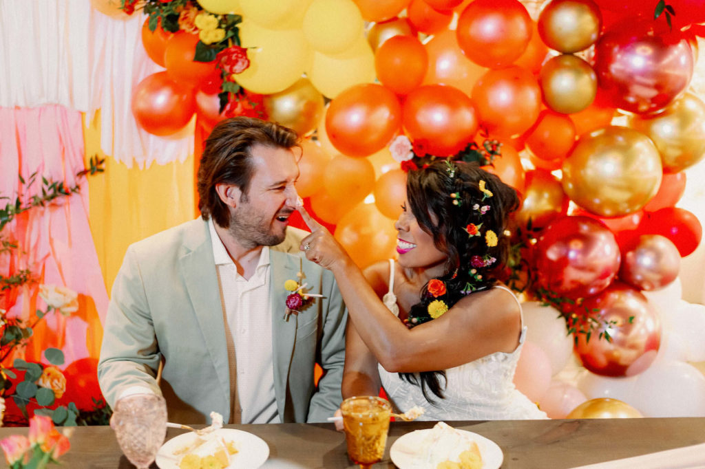 Whimsical Bride with Braided Hair and Flowers and Groom Sitting at Low Picnic Style Table with Neutral Drape Linen, Blush Pink Roses, Tall Candlesticks, Yellow Vintage Glassware, Orange, Yellow, Gold, Pink and Red Balloon Backdrop | Tampa Bay Wedding Photographer Dewitt for Love | Wedding Linen Rentals Kate Ryan Event Rentals | St. Pete Modern Industrial Wedding Venue Red Mesa Events