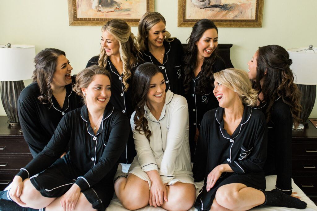 Posh Florida Bridal Party Getting Ready on Bed, Wearing Matching Black Monogram Button Down Pajamas, Bride Wearing All White Pajama Getting Ready PJs