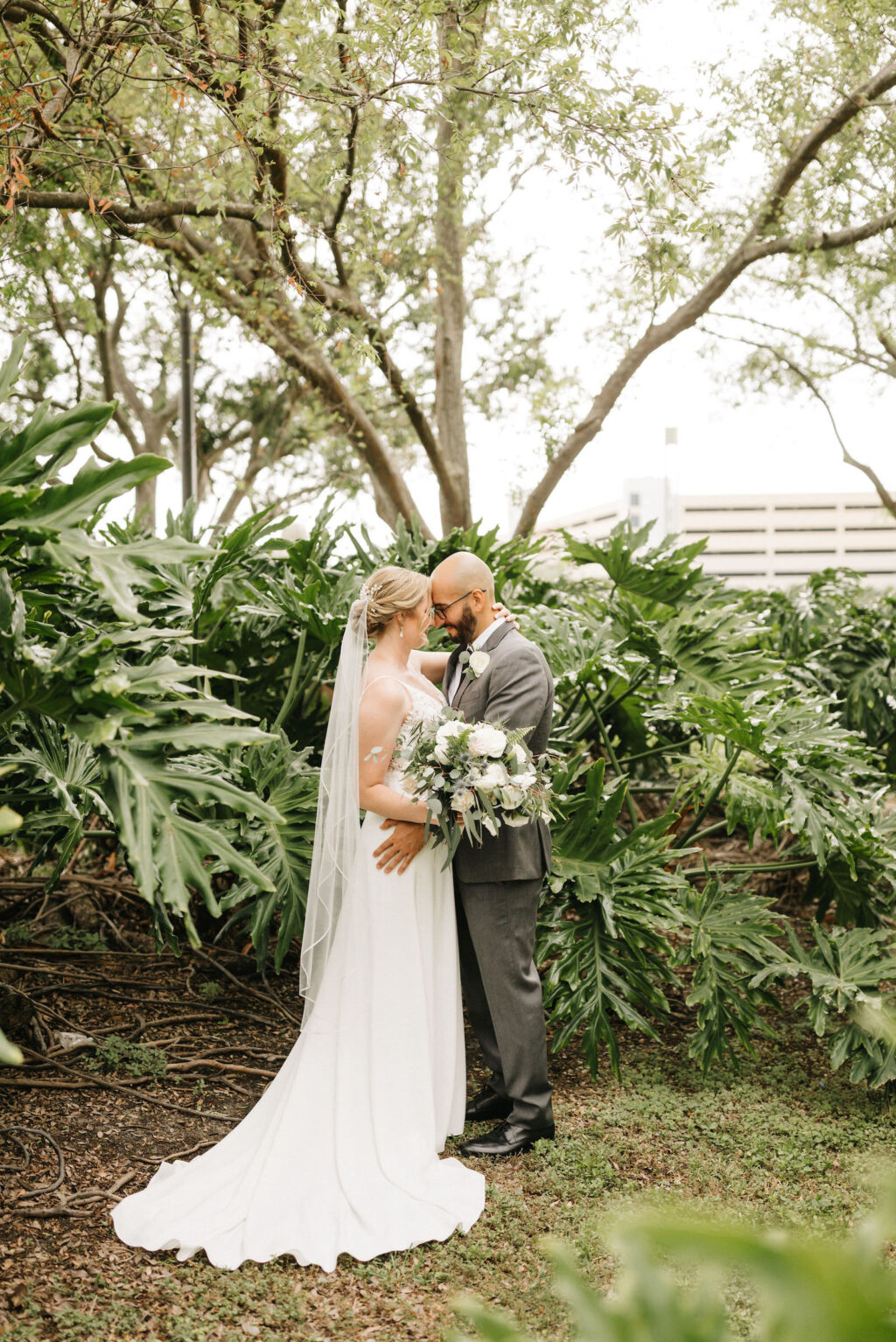 Bride and Groom Outdoor Portrait | Sheath Satin Charmeuse V Neck Lace Bodice Spaghetti Strap Bridal Gown | Groom Wearing Classic Charcoal Grey Suit | Greenery and White Bridal Bouquet
