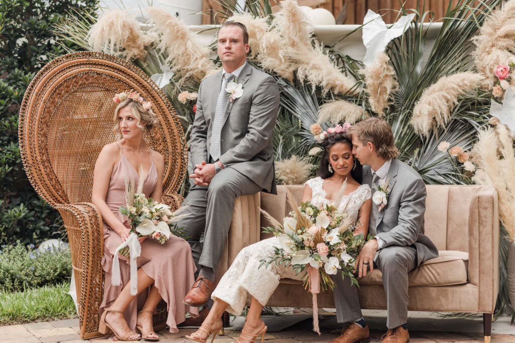Boho, Mid-Century Modern Wedding Decor, Bride Holding Wild Floral Bouquet and Groom on Velvet Cream Loveseat, Bridesmaid Wearing Pink Mauve Dress Sitting in Grand Peacock Rattan Accent Chair, Groomsman Wearing Gray Suit, Pampas Grass and Palm Fronds Backdrop | Tampa Bay Wedding Chair Rentals Kate Ryan Event Rentals | Registration Table Desigbed by Florida Wedding Planner Stephany Perry Events | Big Fake Wedding