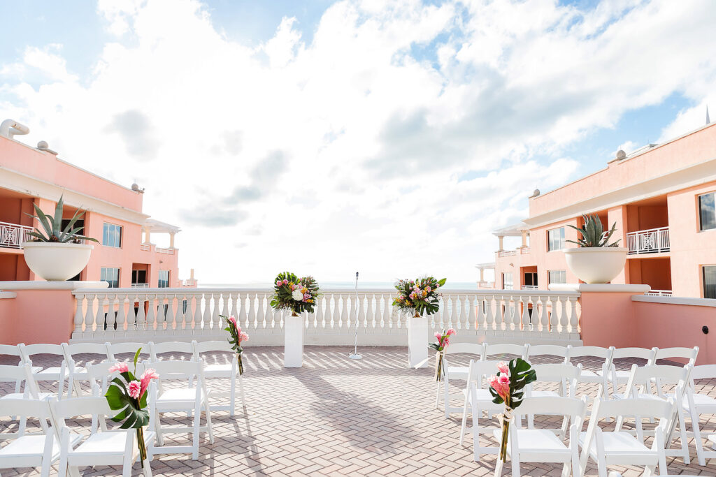 Clearwater Wedding Venue The Hyatt Regency Clearwater Beach | Rooftop Ceremony with White Garden Chairs and White Pillars topped with Bright Pink Tropical Colorful Floral Arrangements and Monstera Palm Leaves | Tampa Bay Wedding Photographer Limelight Photography