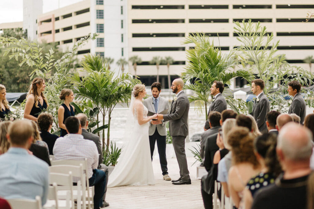 Bride and Groom Exchanging Vows during Waterfront Outdoor Wedding Ceremony in Harbour Island Downtown Tampa | Sheath Satin Charmeuse V Neck Lace Bodice Spaghetti Strap Bridal Gown | Groom Wearing Classic Charcoal Grey Suit