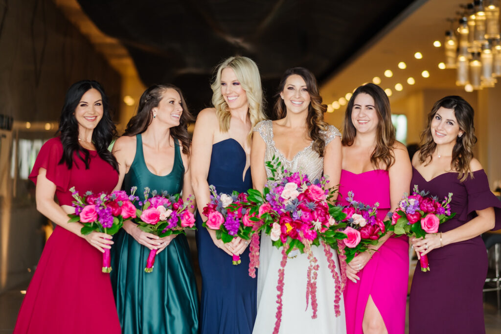 Bride Wearing Gold and Silver Beaded Cap Sleeve Bodice and Flowy Sheath Skirt Wedding Dress Holding Colorful Pink, Purple, Blush Pink, Greenery and Hanging Amaranthus Floral Bouquet, Bridesmaids Wearing Jewel Toned Mismatched Dresses, Red, Turquoise, Blue, Fuschia, and Plum Purple Dresses | Tampa Bay Wedding Florist Iza's Flowers