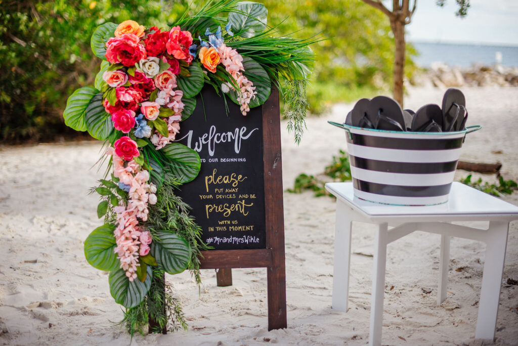 Tropical Jewel Tone Wedding Ceremony Decor, Chalkboard Welcome Sign with Palm Leaves, Pink and Orange Flowers Arrangement, Pail with Black Flip Flops Beach Wedding | Tampa Bay Wedding Florist Iza's Flowers | Wedding Planner Perfecting the Plan