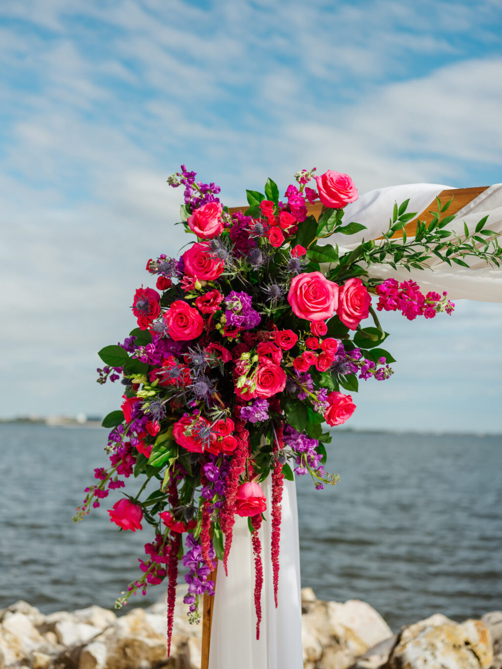 Beach Waterfront Wedding Ceremony Decor, Arch with White Linen Draping, Jewel Tone Pink Roses and Purple Flowers, Hanging Amaranthus Floral Arrangement | Tampa Bay Wedding Planner Perfecting the Plan | Wedding Florist Iza's Flowers | Wedding Venue The Current Hotel Tampa
