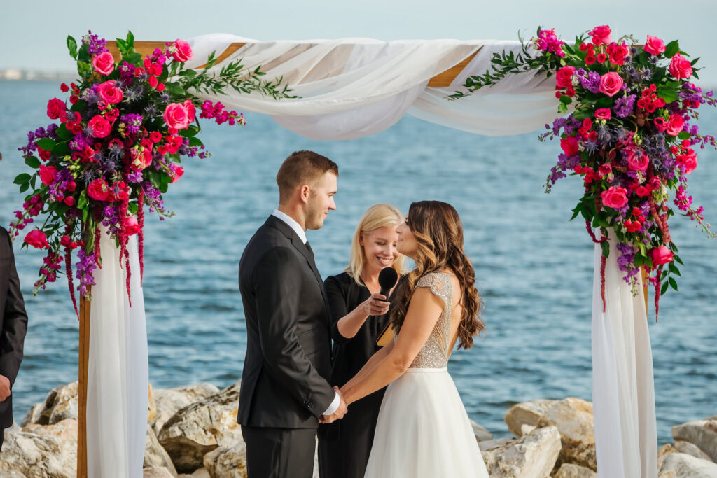Florida Bride and Groom Exchanging Beach Waterfront Wedding Vows Under Arch with White Linen Draping with Jewel Tone Pink Rose, Purple Flowers, Thistle, Hanging Amaranthus and Greenery Floral Arrangements | Tampa Bay Wedding Florist Iza's Flowers | Wedding Planner Perfecting the Plan | Wedding Venue The Current Hotel Tampa | Wedding Officiant Brandi Morris
