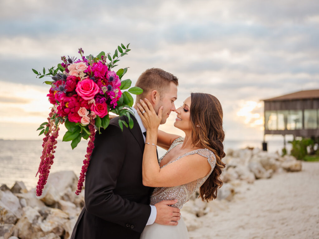 Bride and Groom Kissing on Beach Holding Jewel tone Pink Roses Fuschia Flowers, Thistle, Greenery, Hanging Amaranthus Floral Bouquet | Tampa Bay Wedding Florist Iza's Flowers | Wedding Venue The Current Hotel Tampa