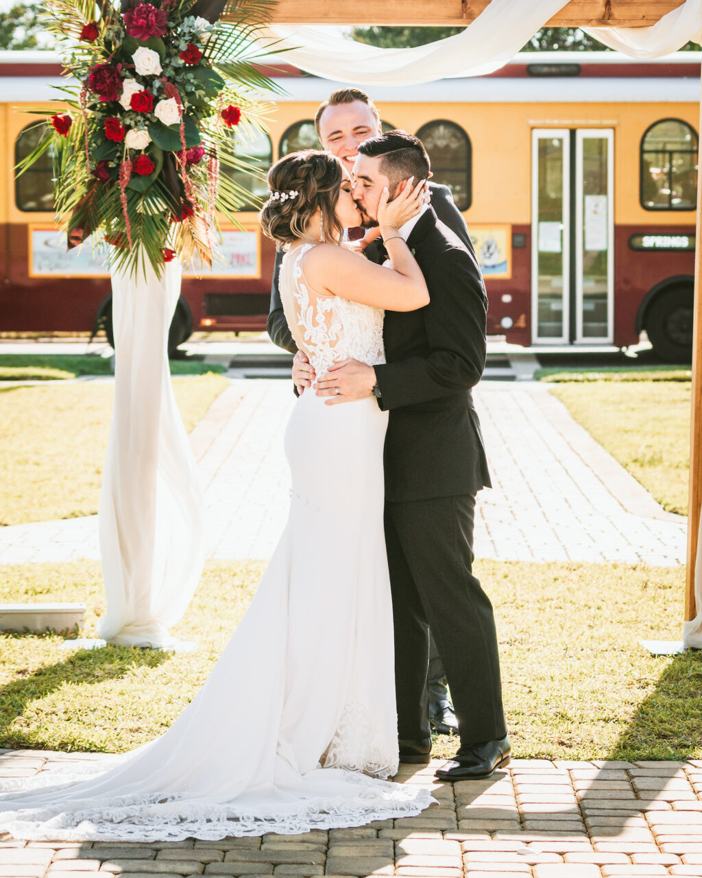 Florida bride and groom exchanging first kiss after exchanging wedding ceremony vows under arch with elegant tropical red roses and palm leaves floral arrangement, tram in background | Tampa Bay wedding photographer Bonnie Newman Creative | Wedding planner Coastal Coordinating | Wedding hair and makeup Femme Akoi | Wedding Florist Iza’s Flowers