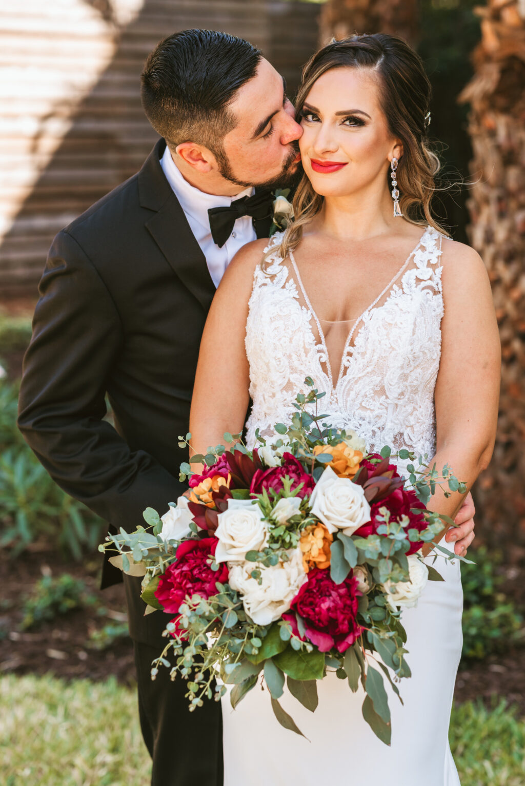 Florida bride wearing floral lace and illusion plunging v neckline wedding dress holding red and white roses, yellow flowers, greenery floral fall bouquet and groom kissing bride on cheek portrait | Tampa Bay wedding photographer Bonnie Newman Creative | Wedding florist Iza’s Flowers Wedding hair and makeup Femme Akoi