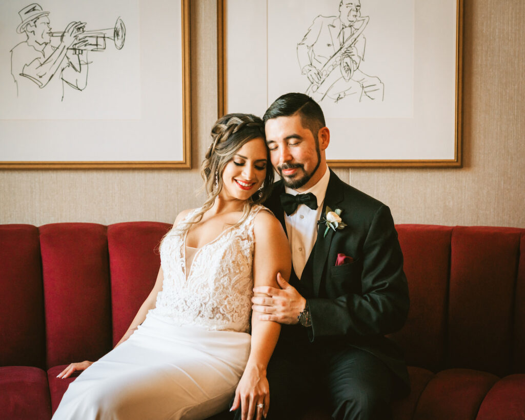 Classic bride and groom sitting on red velvet tufted couch wedding portrait | Tampa Bay wedding photographer Bonnie Newman Creative | Dunedin Wedding Venue Fenway Hotel | Wedding hair and makeup Femme Akoi