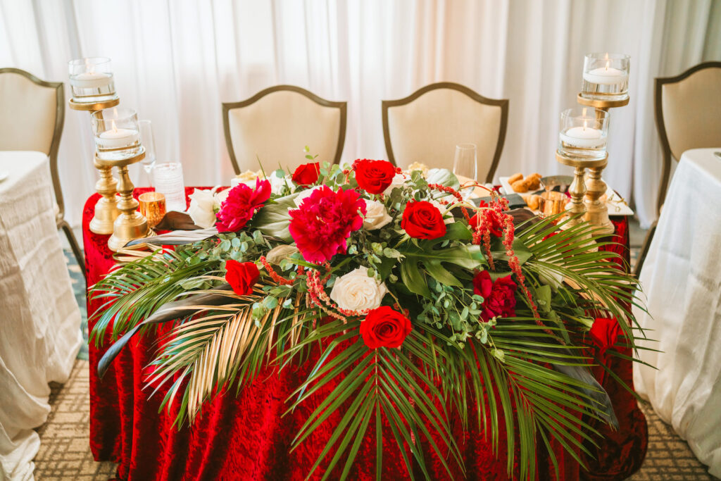 Fall wedding reception decor, sweetheart table with red linen, palm fronds, red and white roses, hanging amaranthus, gold candlestick and floating candles | Tampa Bay wedding photographer Bonnie Newman Creative | Wedding planner Coastal Coordinating | Wedding florist Iza’s Flowers