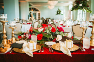 Fall wedding reception decor, sweetheart table with red linen, palm fronds, red and white roses, hanging amaranthus, gold candlestick and floating candles, gold chargers, ivory linen napkin, white favor gift box | Tampa Bay wedding photographer Bonnie Newman Creative | Wedding planner Coastal Coordinating | Wedding florist Iza’s Flowers