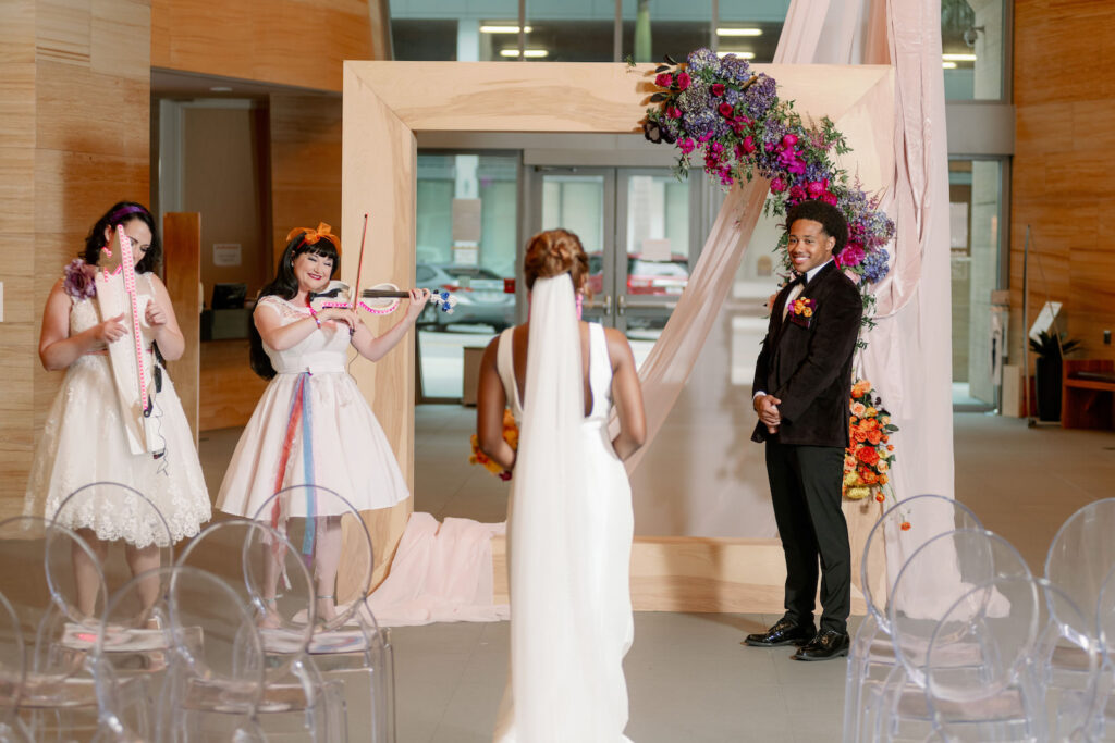 Bride Walking Down Wedding Ceremony Aisle to Groom, Live Musicians, Art Inspired Wedding Ceremony Decor, Wooden Square Frame with White Linen Draping, Whimsical Colorful Cascading Floral Arrangement, Purple, Fuschia Pink, Blush, Orange, Yellow, Acrylic Chairs | Tampa Bay Wedding Planner Wilder Mind Events | St. Pete Wedding Venue The James Museum | Wedding Photographer Dewitt for Love | St. Pete Wedding Venue The James Museum