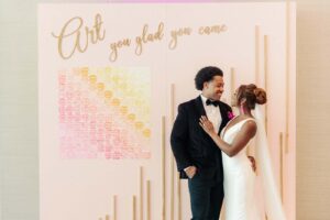 Whimsical Art Inspired Wedding, Modern Bride Wearing Sleek Plunging Neckline Wedding Dress and Pink Tassel Earrings, Groom Wearing Black Tuxedo in front of Bubblegum Pink Wall with Lasercut words "Art you glad you came", Watercolor Pink, Orange and Yellow Seating Chart, Art Gallery at St. Pete Wedding Venue The James Museum | Tampa Wedding Photographer Dewitt for Love