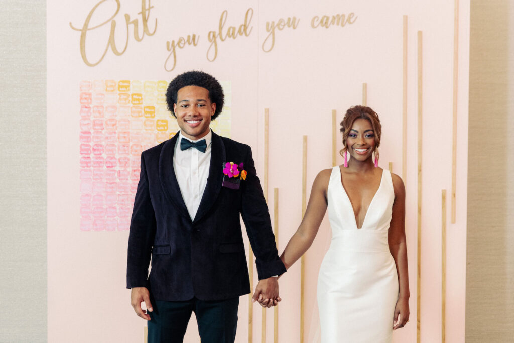 Whimsical Art Inspired Wedding, Modern Bride Wearing Sleek Plunging Neckline Wedding Dress and Pink Tassel Earrings, Groom Wearing Black Tuxedo in front of Bubblegum Pink Wall with Lasercut words "Art you glad you came", Watercolor Pink, Orange and Yellow Seating Chart, Art Gallery at St. Pete Wedding Venue The James Museum | Tampa Wedding Photographer Dewitt for Love