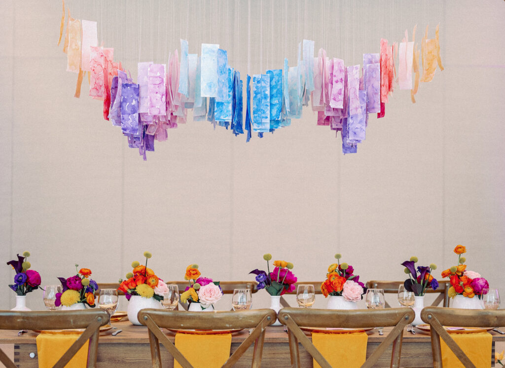 Whimsical Art Inspired Colorful Wedding Reception Decor, Wooden Feasting Table with Wooden Cross Back Chairs, Yellow Linen Napkins, Low Pink, Yellow, Purple, Orang Floral Centerpieces, Hanging Watercolor Papers in Blue, Purple, Pink, Red and Yellow | Tampa Bay Wedding Planner Wilder Mind Events | Wedding Photographer Dewitt for Love | Wedding Rentals Gabro Event Services | Wedding Linens Over the Top Rental Linens | St. Pete Wedding Venue The James Museum