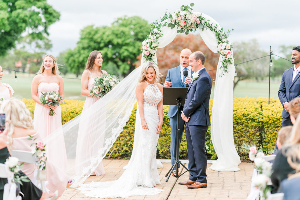Romantic Pink St. Pete Outdoor Garden Wedding Ceremony | Bride and Groom Exchanging Wedding Vows Under String Lights, Round Arch with White Linen Draping and Floral Arrangements | Tampa Bay Wedding Florist Brides N Blooms Designs