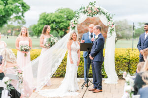 Romantic Pink St. Pete Outdoor Garden Wedding Ceremony | Bride and Groom Exchanging Wedding Vows Under String Lights, Round Arch with White Linen Draping and Floral Arrangements | Tampa Bay Wedding Florist Brides N Blooms Designs