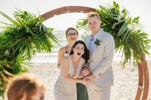 Florida Bride and Groom Reaction During Beachfront Wedding Ceremony, Wooden Circle Arch with Tropical Greenery and Palm Leaves, Groom in Khaki Suit, Bride Wearing Ines Di Santo Wedding Dress