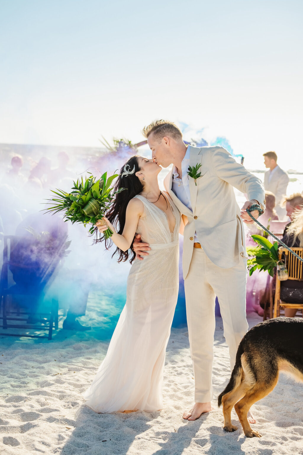 Romantic Florida Bride and Groom Kiss at end of Aisle with Jeweled Toned Smokes Bombs, Bride Wearing Mermaid Style Ines Di Santo Sheer Wedding Dress, Holding Greenery Palm Leaf Bouquet, Groom Wearing Khaki Suit and Barefoot in the sand