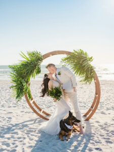 Romantic Florida Bride and Groom Kiss with Pet Dog, in front of Bamboo Circle Arch with Tropical Greenery and Palm Leaf Flora Decor, Bride Wearing Mermaid Style Ines Di Santo Sheer Wedding Dress, Holding Greenery Palm Leaf Bouquet, Groom Wearing Khaki Suit and Barefoot in the sand