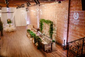 Elegant Wedding Reception and Decor, Exposed Brick Venue with High Ceilings and Vintage lighting, Tropical Inspired Greenery and Floral Arrangements on Long Wooden Feasting Table with Large Square Arch Backdrop | Custom Light Logo | Historic Florida Wedding Venue NOVA 535
