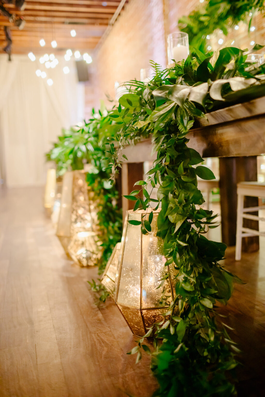 Elegant Wedding Reception and Decor, Exposed Brick with Vintage String lighting, Tropical Inspired Greenery and Floral Arrangements on Long Wooden Feasting Table with Large Square Arch Backdrop | Geometric Mercury Gold Vases | Historic Florida Wedding Venue NOVA 535