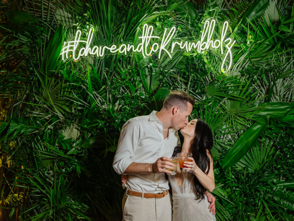 Tampa Bay Bride and Groom Kiss in Front of Photo Backdrop, All Tropical Greenery and Neon Sign with Custom Hashtag