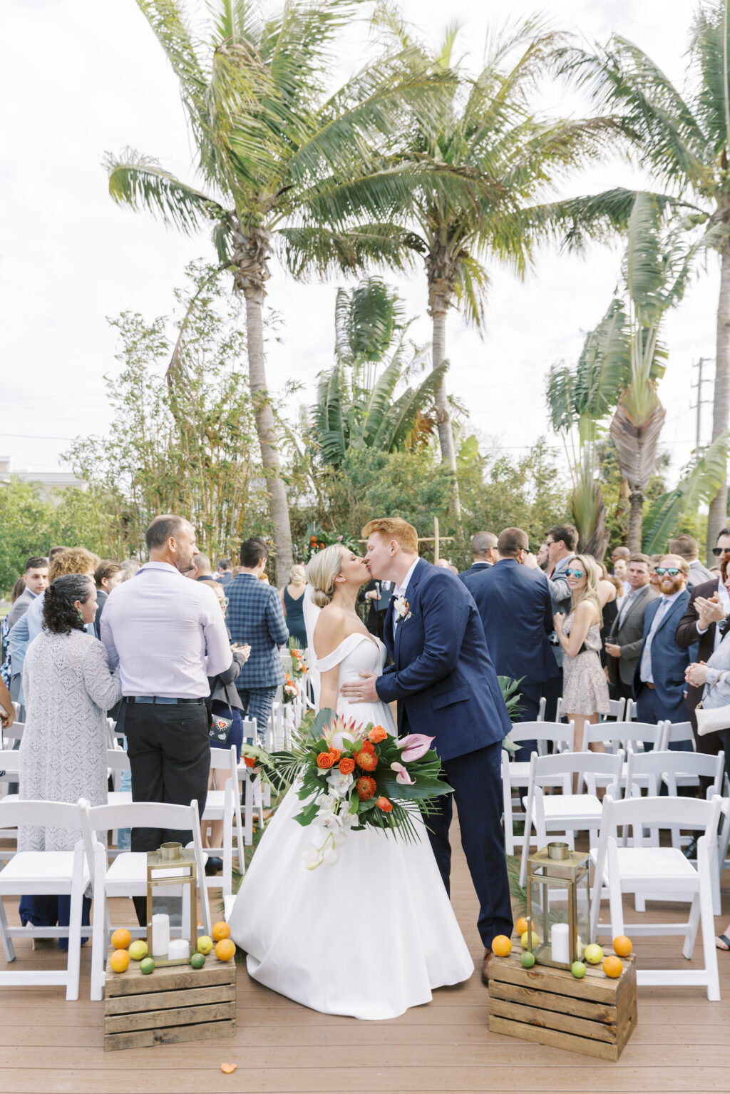 Tropical Pink and Green Wedding Ceremony | Bride and Groom Exchanging Kiss After Wedding Vows | Wooden Pallets with Oranges, Limes, and Lemon Fruits, Gold Lanterns Decor | Tampa Bay Wedding Planner Coastal Coordinating | Outdoor Courtyard St. Pete Wedding Venue Hotel Zamora